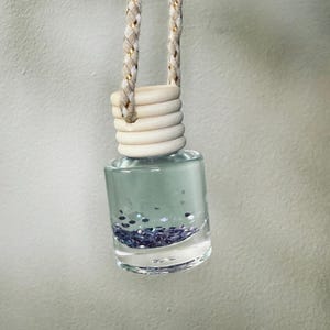 May include: A clear glass car air freshener bottle filled with a light blue liquid and dark blue glitter. The bottle has a wooden cap and is suspended by a braided beige and gold cord.