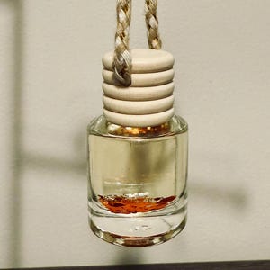 May include: A small, clear glass bottle filled with a light-colored liquid and a reddish-brown substance at the bottom. The bottle has a wooden cap and is suspended by a braided rope. The product is likely an air freshener or fragrance diffuser.