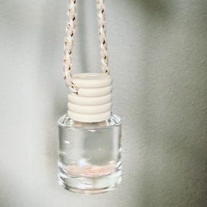 May include: A small, clear glass bottle with a wooden cap and a decorative rope. The bottle contains a pink substance. The rope is white with gold accents. The bottle is designed to hang.