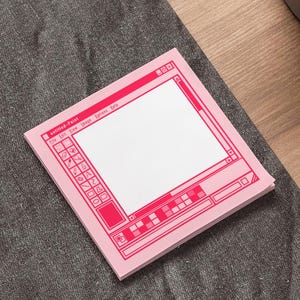 May include: A square sticky note pad featuring a bright pink graphic print of a classic computer paint software interface. The design includes a toolbar on the left, a large central white workspace, and menu bars at the top and bottom of the frame.