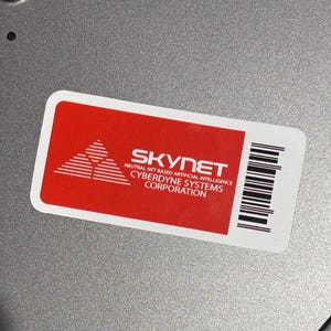 Può includere: Un adesivo rosso e bianco con la scritta "SKYNET" e "NEURAL NET BASED ARTIFICIAL INTELLIGENCE CYBERDYNE SYSTEMS CORPORATION" è attaccato a una superficie argentata. È presente anche uno stilo bianco.