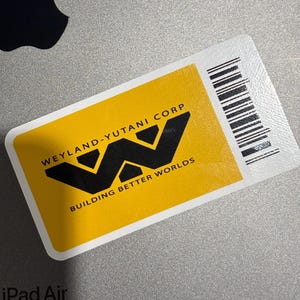 May include: A yellow and black sticker with the text "WEYLAND-YUTANI CORP BUILDING BETTER WORLDS" is placed on a gray iPad Air. A white stylus and the Apple logo are also visible.