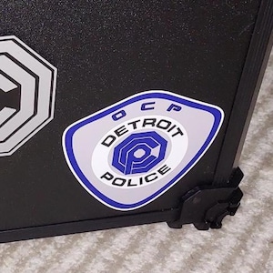 Puede incluir: Maleta negra de carcasa dura con varias pegatinas. Las pegatinas incluyen un diseño geométrico blanco y gris, una insignia de la policía de Detroit azul y blanca y una pegatina con las letras "HMF".