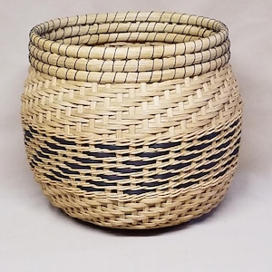 May include: A handwoven basket with a round shape. The basket is made of natural-colored reeds with a dark blue band around the middle. The rim is reinforced with multiple layers of the same material.