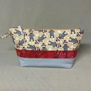 May include: A rectangular fabric pouch with a zipper closure. The top section features a beige background with sock monkey patterns and buttons. A red patterned band separates it from a light blue base. A small fabric loop is attached to the zipper.