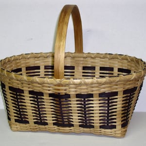 May include: A woven basket with a brown wooden handle. The basket is made of natural materials and has a black and brown checkered pattern.