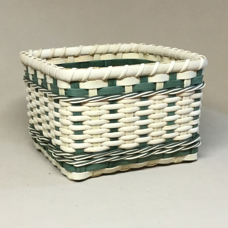 Peut inclure: Un panier carr&eacute; tiss&eacute;, fabriqu&eacute; avec des roseaux blancs et verts. Le panier pr&eacute;sente un motif tiss&eacute; et une bordure verte autour du haut.