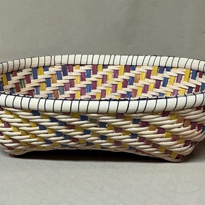 May include: A woven basket with an oval shape. The basket is made of natural-colored reeds and features a colorful pattern of yellow, blue, and purple. The rim is white with black stripes.