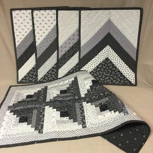 May include: Set of five black and white quilted placemats. The placemats feature a geometric pattern of black and white triangles and squares. The placemats are made of fabric and are perfect for adding a touch of style to any dining table.