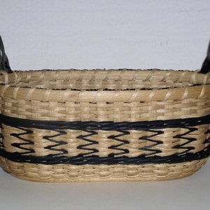 May include: A woven basket with a rectangular shape and black leather handles. The basket features a natural tan color with a black zigzag pattern around the middle. The handles are attached to the top edges.