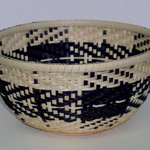 May include: A woven basket with a black and white geometric pattern. The basket is made of natural materials and has a round shape.