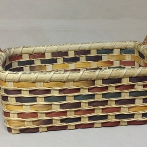 May include: A rectangular woven basket with leather handles. The basket is made of natural colored, brown, red, and green woven reeds.