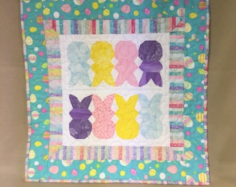 May include: A colorful patchwork quilt with a border of pastel blue and green fabric with white polka dots and a pattern of Easter eggs. The center of the quilt features a design of six pastel-colored bunnies.