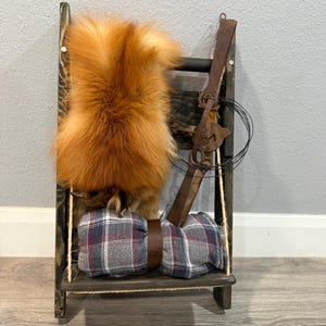May include: A rustic wooden shelf displays a vibrant orange fox pelt, a vintage metal trap, and two plaid fabric bundles secured with a brown leather strap. The shelf is suspended by rope.
