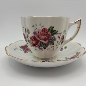 May include: White teacup and saucer set with a floral rose design in shades of pink and green. The cup and saucer have a scalloped edge and gold trim. The teacup handle is also trimmed in gold.