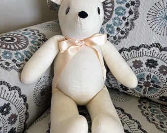May include: A cream-colored stuffed animal mouse with a black nose and small black eyes. The mouse has large, rounded ears and a light pink ribbon tied in a bow around its neck. The toy is sitting on a patterned chair.