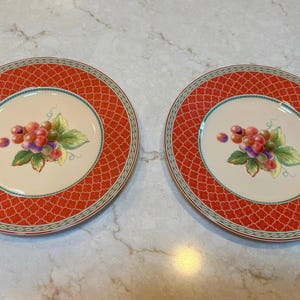 May include: Two round decorative plates. Each plate has a red patterned border and a cream-colored center. The center of each plate features a cluster of grapes with green leaves.