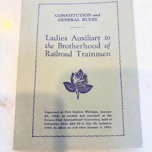 May include: A vintage book cover with the title "CONSTITUTION and GENERAL RULES" and "Ladies Auxiliary to the Brotherhood of Railroad Trainmen" in dark blue ink. A rose illustration is centered below the title. The book is light blue.