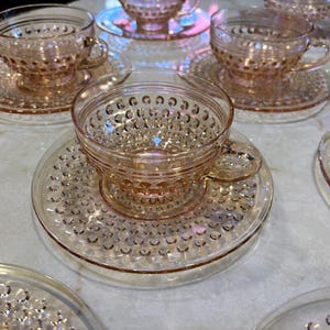 May include: A collection of vintage pink glass teacups and saucers. The cups and saucers have a textured, hobnail design. The set is arranged on a light-colored surface, suggesting a tea party or display of antique glassware.