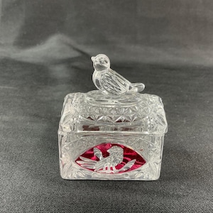 May include: A clear, cut-glass trinket box with a bird figurine on top. The box has a square shape with a decorative pattern and a red interior. A bird is etched on the front.