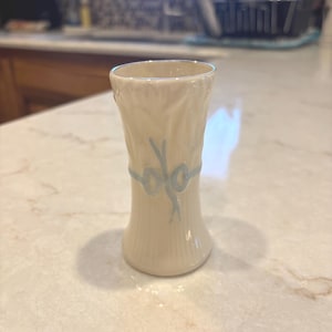 May include: A small, cream-colored ceramic vase with a vertical ribbed texture. The vase features a light blue painted design around the center, resembling a tied bundle of wheat or stalks, with a flared top rim and a slightly wider base.