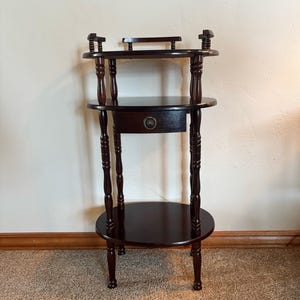 May include: A dark brown wooden tiered stand featuring three circular shelves, four turned spindle legs, and a single central drawer with a round metal pull. The top shelf includes a small raised gallery rail with decorative spindle accents.