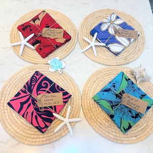 May include: Four square fabric napkins with tropical patterns in red, blue, and pink, each tied with twine and a brown paper tag labeled with names and dimensions in inches. The napkins rest on round woven placemats decorated with white starfish.