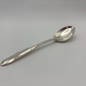 May include: A silver spoon with a rounded bowl and a long, slender handle. The handle features a delicate floral design. The spoon is set against a plain, light gray background.