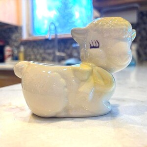 May include: A cream-colored ceramic planter shaped like a resting lamb. The lamb features a glossy finish, a sculpted bow around its neck, and painted closed eyes with long eyelashes. The opening for plants is located on the lamb's back.