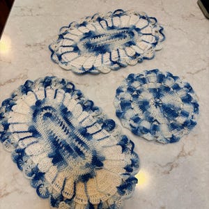 May include: Three handmade crochet doilies in varying sizes. The doilies feature a white and blue color scheme with an oval and circular shape. The edges have a scalloped design. The doilies are made of a soft, textured yarn.