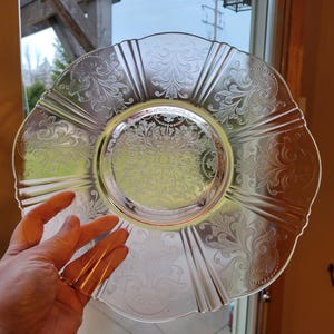 May include: Clear glass plate with a scalloped edge and intricate floral pattern. The plate has a central design and radiating lines. A hand is holding the plate in front of a window.