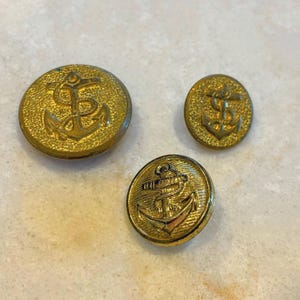 May include: Three round gold-toned metal buttons of varying sizes, each featuring a raised anchor emblem. The two larger buttons have a textured, pebbled background, while the smaller button displays a detailed anchor with a rope design.