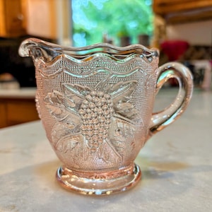 May include: Pink glass creamer with a detailed grape and leaf design. The creamer has a handle and a scalloped rim. The glass has a textured surface and sits on a round base. A vintage-style piece.