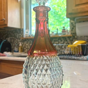May include: A glass decanter featuring a clear, diamond-patterned base that transitions into a smooth, iridescent red neck. The decanter includes a matching clear, faceted, diamond-shaped glass stopper that sits securely in the narrow opening.