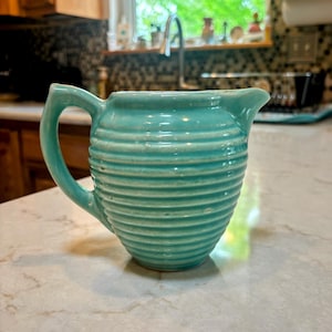 May include: A turquoise ceramic pitcher with a ribbed design. The pitcher has a curved handle and a spout for pouring. The kitchenware is sitting on a white countertop.