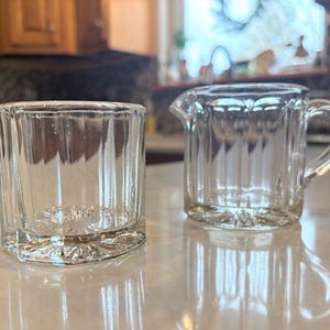 May include: Clear glass creamer and tumbler set. The creamer has a handle and spout, while the tumbler is a short, square shape. Both pieces have vertical ribbed detailing and decorative bases, reflecting light.