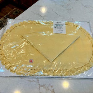 May include: A new, packaged yellow oval placemat and matching napkin set. The placemat has a decorative lace edge. The set is sealed in clear plastic and includes a product information tag.