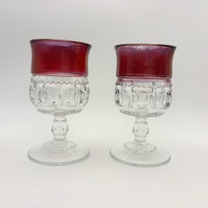 May include: Two clear glass goblets with a red rim. The goblets have a decorative pattern on the bowl and a stem with a round base. The red rim adds a pop of color to the otherwise transparent glass.