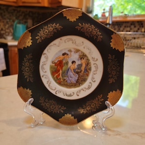 May include: A decorative black plate with gold floral accents and a central image of figures in classical attire. The plate is displayed on a clear plastic stand, showcasing its ornate design and vintage aesthetic.