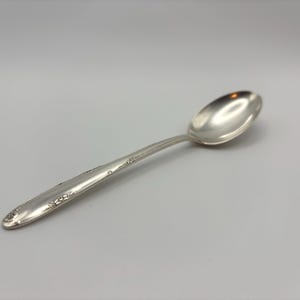 May include: A polished silver-toned metal spoon featuring an elongated oval bowl and a slender handle. The handle displays a subtle, raised floral relief pattern along its edges, extending from the base toward the neck of the spoon.