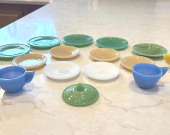 May include: A collection of miniature toy dishes including several small cups, saucers, and a single lid. The pieces are made of solid-colored plastic in shades of green, yellow, blue, white, and tan, arranged in a scattered group on a surface.