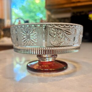 May include: A clear glass pedestal bowl with intricate floral and geometric patterns. The bowl sits on a red-tinted base. The design is visible on the bowl's exterior, adding a decorative element to the piece.