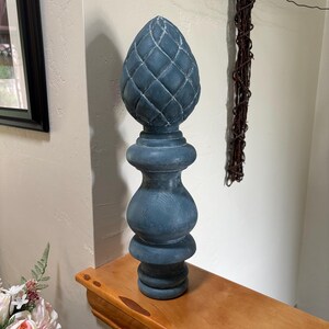 May include: A decorative finial featuring a blue, distressed finish with a pinecone-shaped top and a multi-tiered, rounded pedestal base. The surface shows visible wood grain texture throughout the structure, highlighting the carved details.
