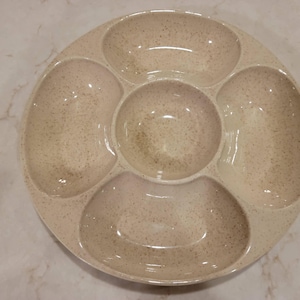 May include: A speckled beige ceramic serving platter with five compartments. The round platter has a central circular bowl surrounded by four larger, curved sections. The surface has a speckled pattern.