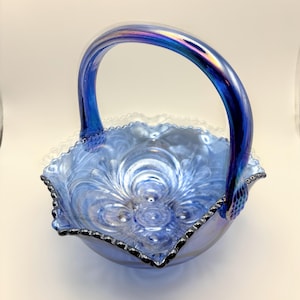 May include: A blue glass basket with a handle. The basket has a ruffled edge and a floral pattern on the inside. The handle is arched and has an iridescent sheen. The basket is likely vintage and suitable for display or decorative use.