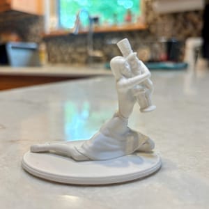 May include: A white porcelain figurine depicting a woman embracing a man in a top hat. The sculpture is mounted on a round, white base. The woman is kneeling, and the man is standing. The figurine is a decorative item.