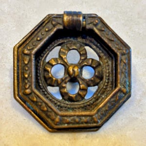 May include: An antique bronze octagonal drawer pull with a floral center design. The pull has an ornate, detailed border and a small handle at the top. The metal has a weathered appearance, with hints of green and blue oxidation.