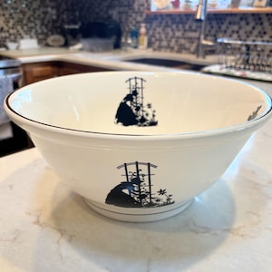 May include: A white ceramic bowl with a black rim and a decorative black silhouette design. The design features a figure tending a garden trellis. The bowl is on a light-colored surface.