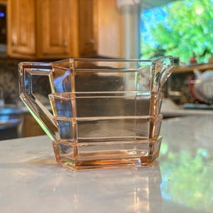 May include: A vintage, pink glass creamer with a geometric design. The creamer has a rectangular handle and a spout for easy pouring. The glass is a soft, translucent pink, and the creamer is sitting on a white surface.