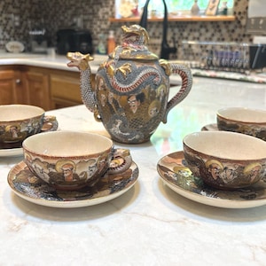 May include: A ceramic tea set featuring a teapot with a dragon-shaped spout and lid handle, accompanied by three matching teacups and saucers. The set is decorated with intricate gold, black, and multi-colored illustrations of figures and dragons.